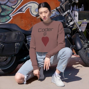 Może przedstawiać: Kobieta w brązowej, krótkiej bluzie z napisem "Coder @" i czerwonym sercem. Siedzi przed motocyklem.