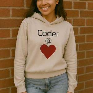 May include: A beige hoodie with the word "Coder" above a red heart graphic. The "@" symbol is between the words and the heart. The person is wearing blue jeans and is leaning against a brick wall.