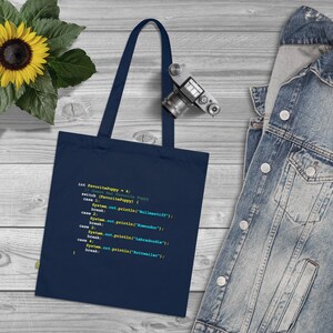 May include: A navy blue tote bag with a code snippet printed on it. The code is written in Java and is a simple switch statement that prints the name of a dog breed based on a number. The code is written in a light blue font with yellow highlights.