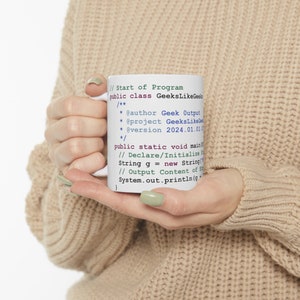 Könnte beinhalten: Eine weiße Keramiktasse mit einem grün-blauen Design, das Code aus einem Programm namens "GeeksLikeGeeks" zeigt. Der Code enthält Kommentare, eine Klassendefinition und eine Hauptmethode.