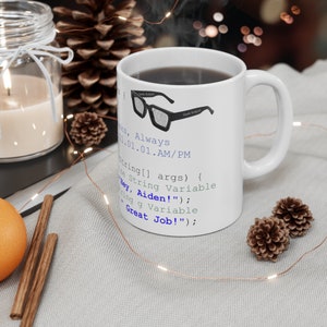 May include: White ceramic coffee mug with a blue and black design featuring code and a pair of glasses. The text on the mug reads "s, Always 1.01.01.AM/PM String[] args) { ze String Variable Bey, Aiden!" ; ting q Variable Great Job!" ;"