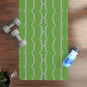 May include: A green yoga mat with a repeating gray geometric pattern. Two gray dumbbells and a water bottle with blue liquid are on the wooden floor next to the mat. A small potted plant is also visible.