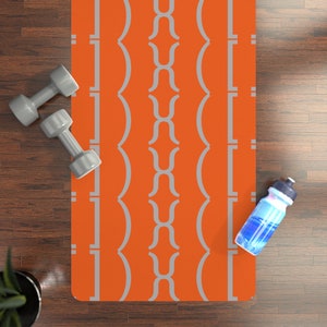 May include: An orange yoga mat with a gray geometric pattern is displayed on a wooden floor. Two gray dumbbells and a water bottle are also present. The mat is rectangular and features a repeating design. The water bottle is blue and white.