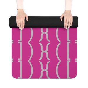 May include: A rolled-up yoga mat with a black top and a vibrant pink surface. The pink surface features a repeating gray geometric pattern. The mat is held by two hands with light skin.