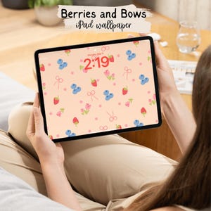 Puede incluir: Una tableta digital que muestra un fondo de pantalla rosa y blanco con un patrón de fresas rojas, arándanos azules, flores rosas y lazos blancos. La hora es 2:19 en la pantalla.