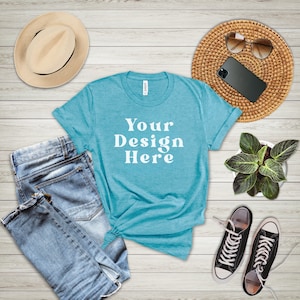May include: A turquoise blue t-shirt with the text "Your Design Here" in white. The t-shirt is laid out on a white wood background with blue jeans, a straw hat, sunglasses, a potted plant, and black sneakers.