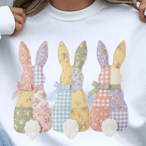 Peut inclure: Sweat-shirt blanc orné de trois lapins appliqués. Chaque lapin présente un motif patchwork avec des motifs floraux, vichy et unis aux couleurs pastel. Les lapins ont des nœuds et des accents de queue blancs texturés.