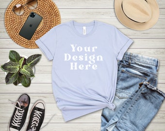 May include: A light blue t-shirt with the text "Your Design Here" in white. The t-shirt is laid flat on a wooden surface with a pair of black sneakers, a straw hat, a woven coaster, and a potted plant.