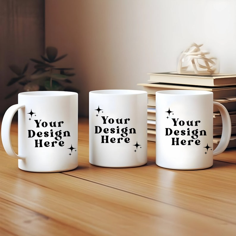 May include: Three white ceramic mugs with black text that reads "Your Design Here" and small black star designs. The mugs are on a wooden surface.