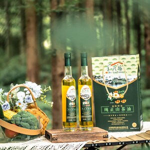 May include: Two glass bottles of green tea oil with a green and white box labelled "真山茶油" in Chinese characters. The bottles are on a wooden table with a basket of vegetables and flowers, corn on the cob, and tomatoes.