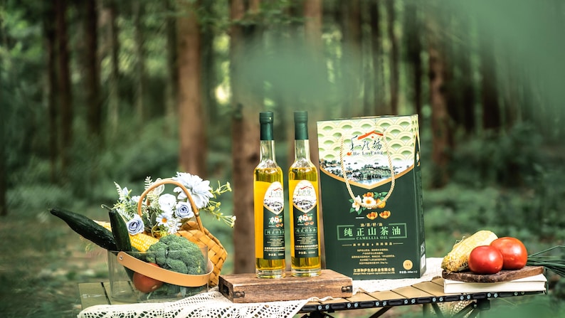 May include: Two glass bottles of green tea oil with a green and white box labelled "真山茶油" in Chinese characters. The bottles are on a wooden table with a basket of vegetables and flowers, corn on the cob, and tomatoes.