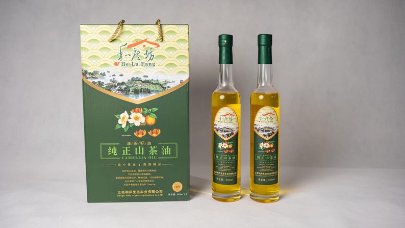 May include: Two glass bottles of camellia oil with green labels and a green box with Chinese text and a picture of a camellia flower. The text on the label reads "Camellia Oil".