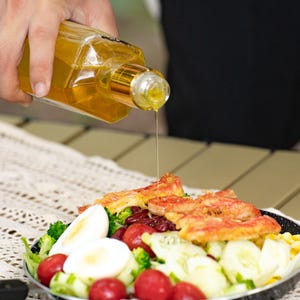 May include: A black pan filled with a salad of broccoli, cherry tomatoes, cucumber, red beans, hard-boiled eggs, and crab meat. A person is pouring oil from a clear bottle over the salad.