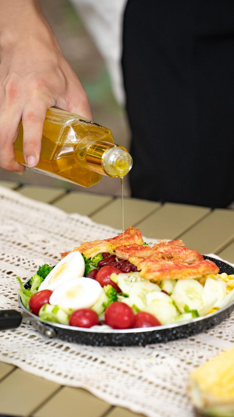 May include: A black pan filled with a salad of broccoli, cherry tomatoes, cucumber, red beans, hard-boiled eggs, and crab meat. A person is pouring oil from a clear bottle over the salad.