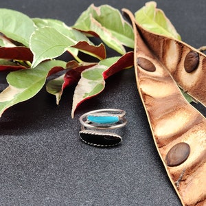 May include: A silver ring with a black and turquoise stone. The ring is wrapped around a finger and has a delicate design.