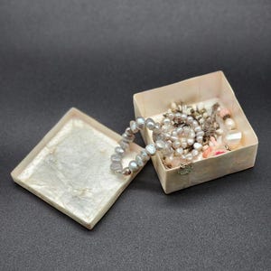 Puede incluir: Una caja blanca cuadrada texturizada con una tapa abierta, que revela una colección de collares de perlas y otras joyas. La caja está sobre una superficie gris oscuro.