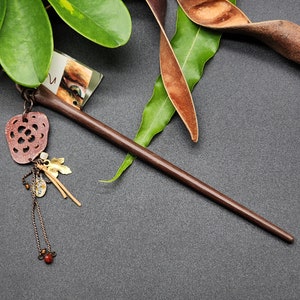 May include: A dark brown wooden hair stick with decorative charms. The stick is long and slender, with a carved wooden charm, small gold-coloured leaves, and a beaded chain with a red bead. The background includes green leaves and a dark surface.