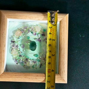 Pu&ograve; includere: Un'opera d'arte incorniciata con una pietra verde centrale circondata da un motivo a spirale e varie pietre preziose. La cornice &egrave; in legno chiaro. L'opera misura circa 7,6 cm di altezza.