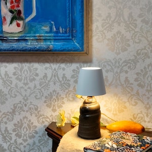 May include: A small, dark brown ceramic lamp with a gray lampshade sits on a wooden table. A painting with a blue frame hangs on the wall. A stack of patterned notebooks and a yellow flower are on the table. A piece of fruit is also on the table.