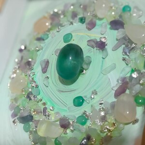 Pu&ograve; includere: Primo piano di un elemento decorativo con una grande pietra verde lucida al centro, circondata da pietre pi&ugrave; piccole nei toni del verde, viola e bianco. Scaglie d'argento aggiungono brillantezza allo sfondo dipinto di verde chiaro.