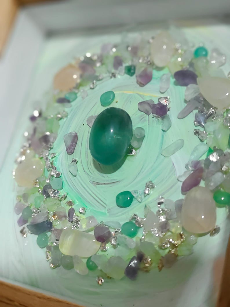Pu&ograve; includere: Primo piano di un elemento decorativo con una grande pietra verde lucida al centro, circondata da pietre pi&ugrave; piccole nei toni del verde, viola e bianco. Scaglie d'argento aggiungono brillantezza allo sfondo dipinto di verde chiaro.