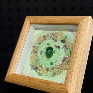 Pu&ograve; includere: Opera d'arte incorniciata con un design circolare di pietre preziose. L'opera presenta uno sfondo verde chiaro, una pietra verde centrale e un bordo di varie pietre preziose. La cornice &egrave; in legno chiaro.
