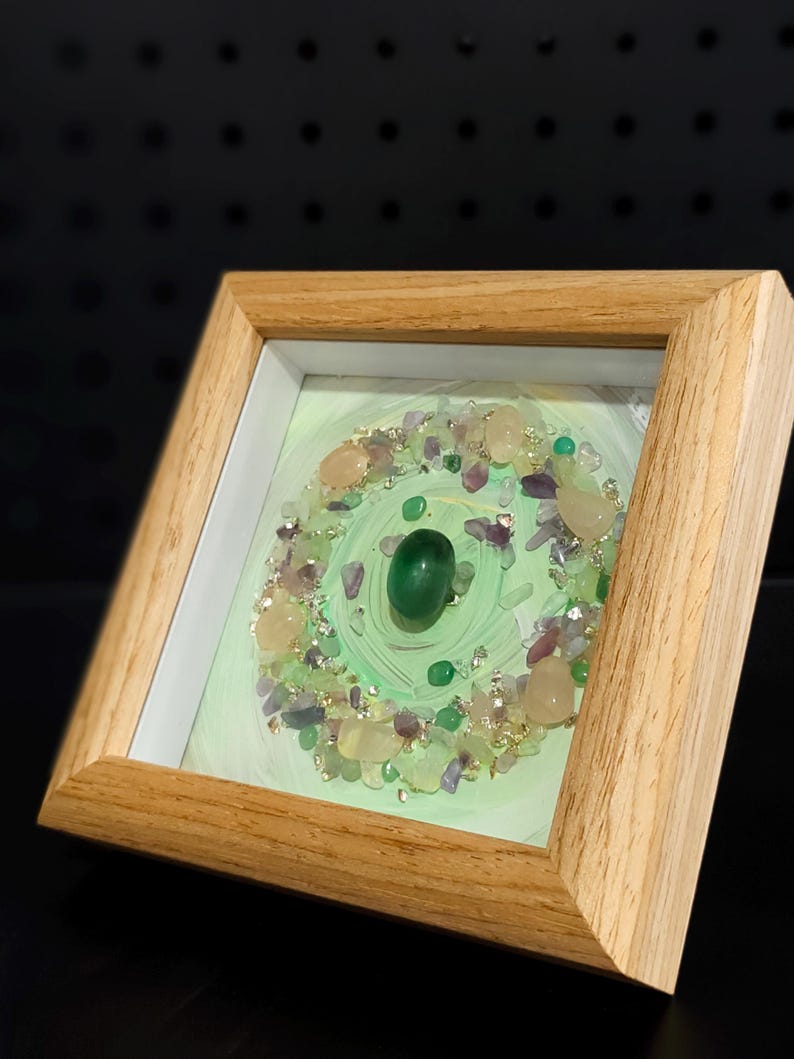 Pu&ograve; includere: Opera d'arte incorniciata con un design circolare di pietre preziose. L'opera presenta uno sfondo verde chiaro, una pietra verde centrale e un bordo di varie pietre preziose. La cornice &egrave; in legno chiaro.