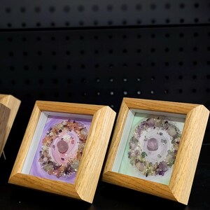 Pu&ograve; includere: Due opere d'arte incorniciate con una disposizione circolare di cristalli e pietre colorate. L'opera d'arte &egrave; ambientata su uno sfondo dipinto con tonalit&agrave; viola e verdi. Le cornici sono in legno chiaro.