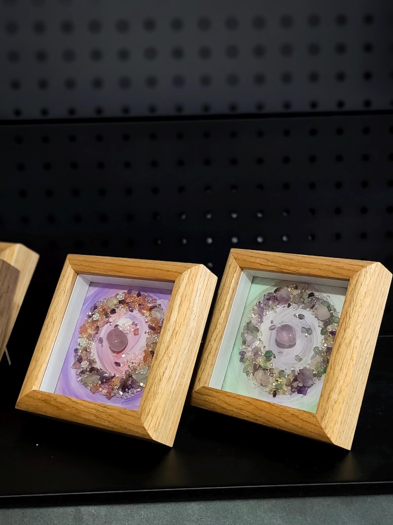 Pu&ograve; includere: Due opere d'arte incorniciate con una disposizione circolare di cristalli e pietre colorate. L'opera d'arte &egrave; ambientata su uno sfondo dipinto con tonalit&agrave; viola e verdi. Le cornici sono in legno chiaro.