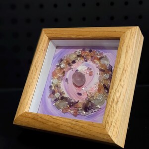 Pu&ograve; includere: Un'opera d'arte incorniciata con una disposizione circolare di cristalli di ametista, quarzo rosa e quarzo trasparente. I cristalli sono disposti su uno sfondo di vernice viola e rosa a spirale, all'interno di una cornice di legno chiaro.