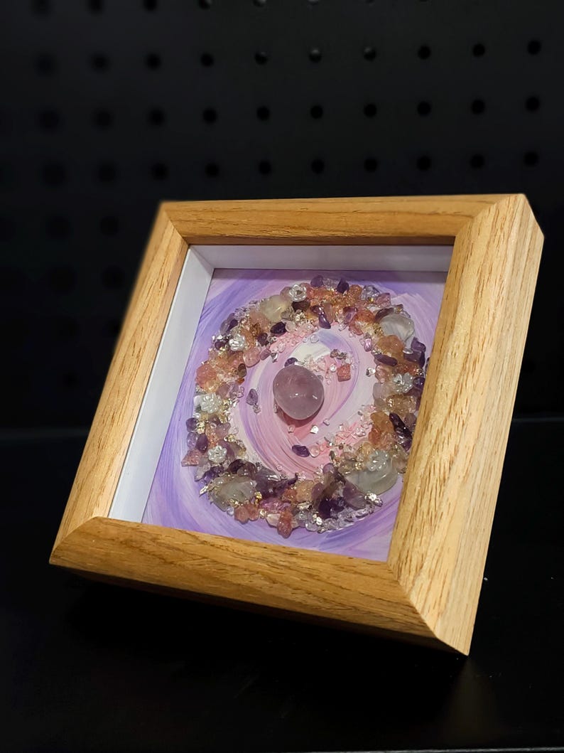 Pu&ograve; includere: Un'opera d'arte incorniciata con una disposizione circolare di cristalli di ametista, quarzo rosa e quarzo trasparente. I cristalli sono disposti su uno sfondo di vernice viola e rosa a spirale, all'interno di una cornice di legno chiaro.