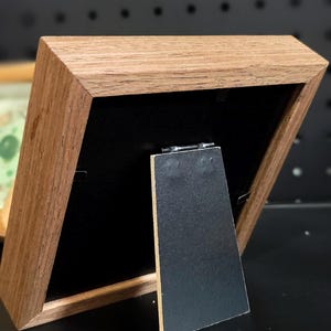 Pu&ograve; includere: Una piccola cornice in legno con finitura a venatura naturale. La cornice ha un retro nero e un cavalletto nero. La cornice &egrave; angolata, mostrando il retro e il profilo laterale. La cornice &egrave; appoggiata su una superficie nera.