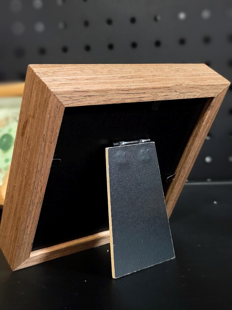 Pu&ograve; includere: Una piccola cornice in legno con finitura a venatura naturale. La cornice ha un retro nero e un cavalletto nero. La cornice &egrave; angolata, mostrando il retro e il profilo laterale. La cornice &egrave; appoggiata su una superficie nera.