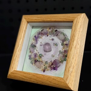 Pu&ograve; includere: Opera d'arte incorniciata con una disposizione circolare di ametista, quarzo e cristalli di fluorite verde. I cristalli sono disposti su uno sfondo dipinto di verde chiaro e bianco, all'interno di una cornice in legno naturale.