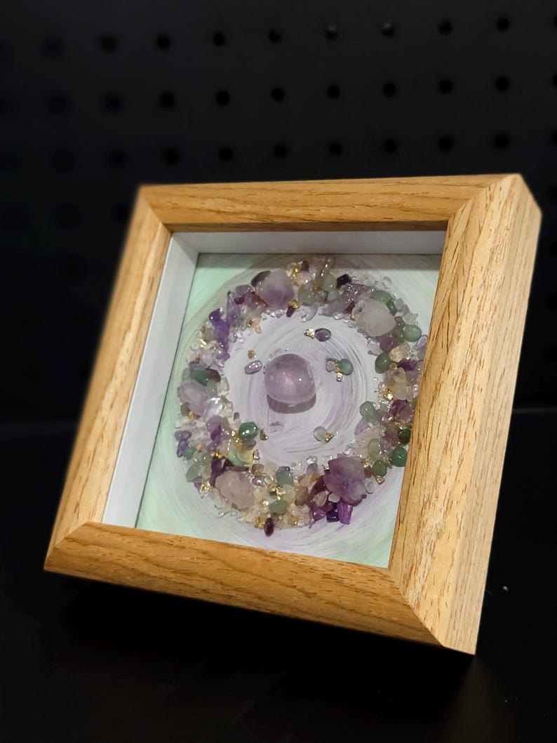 Pu&ograve; includere: Opera d'arte incorniciata con una disposizione circolare di ametista, quarzo e cristalli di fluorite verde. I cristalli sono disposti su uno sfondo dipinto di verde chiaro e bianco, all'interno di una cornice in legno naturale.