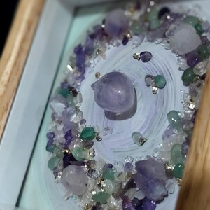 Pu&ograve; includere: Un'opera d'arte incorniciata con una disposizione centrale di cristalli di ametista e quarzo lucidi, circondata da un mix di piccole pietre preziose colorate. Lo sfondo presenta un motivo dipinto in blu chiaro e viola, con una cornice in legno naturale.