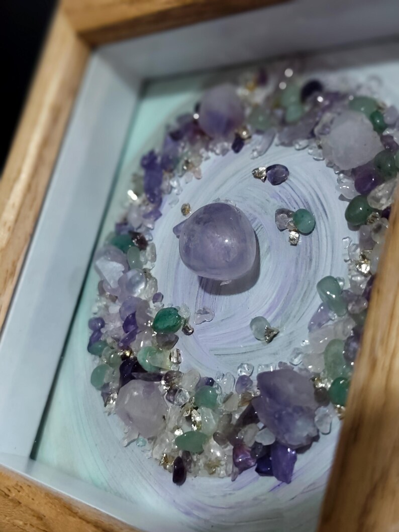Pu&ograve; includere: Un'opera d'arte incorniciata con una disposizione centrale di cristalli di ametista e quarzo lucidi, circondata da un mix di piccole pietre preziose colorate. Lo sfondo presenta un motivo dipinto in blu chiaro e viola, con una cornice in legno naturale.