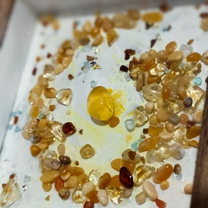 Pu&ograve; includere: Una collezione di pietre preziose lucide nelle tonalit&agrave; del giallo e dell'arancione, disposte su una superficie bianca all'interno di una cornice di legno. Le pietre preziose variano per dimensioni e forma, creando un effetto strutturato. Una grande gemma gialla rotonda &egrave; al centro.