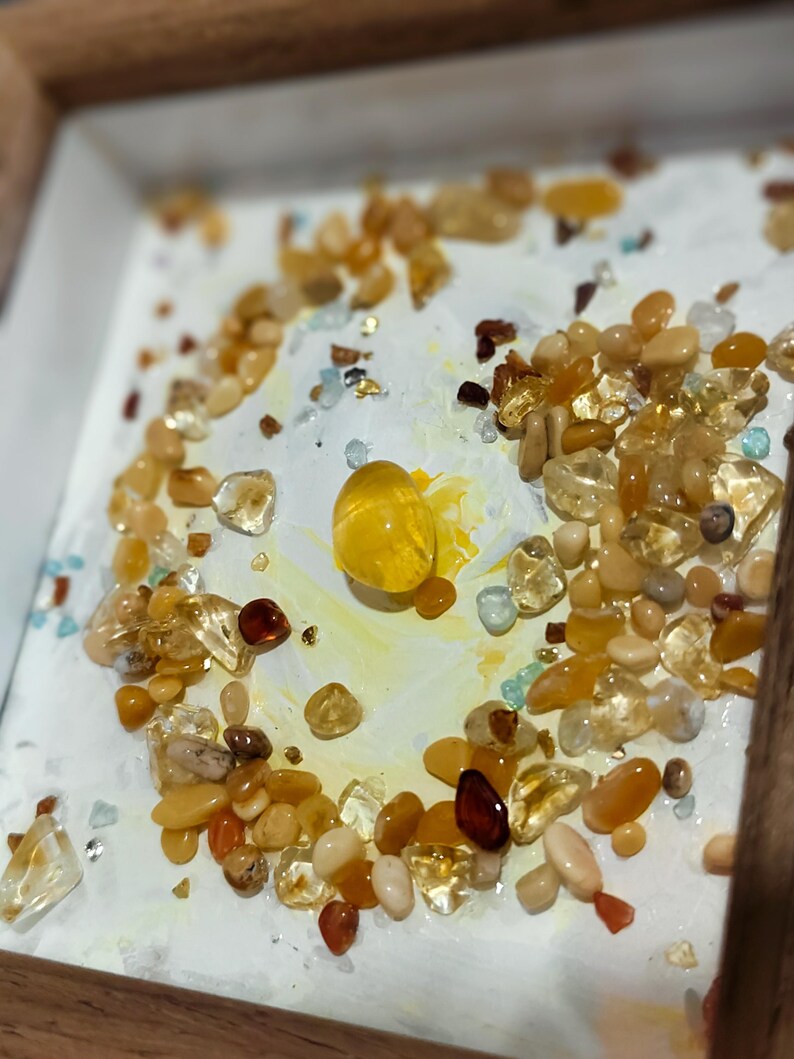 Pu&ograve; includere: Una collezione di pietre preziose lucide nelle tonalit&agrave; del giallo e dell'arancione, disposte su una superficie bianca all'interno di una cornice di legno. Le pietre preziose variano per dimensioni e forma, creando un effetto strutturato. Una grande gemma gialla rotonda &egrave; al centro.