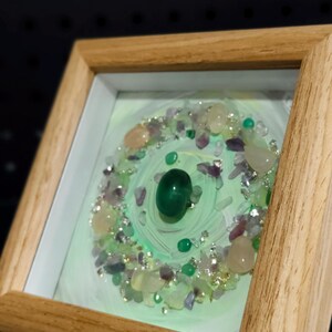 Pu&ograve; includere: Opera d'arte incorniciata con una gemma verde centrale, circondata da pietre pi&ugrave; piccole nei toni del verde, viola e crema. L'opera &egrave; ambientata su uno sfondo verde chiaro e racchiusa in una cornice di legno naturale.