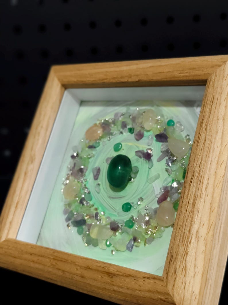 Pu&ograve; includere: Opera d'arte incorniciata con una gemma verde centrale, circondata da pietre pi&ugrave; piccole nei toni del verde, viola e crema. L'opera &egrave; ambientata su uno sfondo verde chiaro e racchiusa in una cornice di legno naturale.