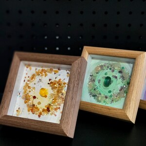 Pu&ograve; includere: Due opere d'arte quadrate incorniciate. Una presenta una pietra gialla centrale circondata da pietre arancioni e gialle su sfondo bianco. L'altra ha una pietra verde centrale con pietre verdi e viola su sfondo verde chiaro.