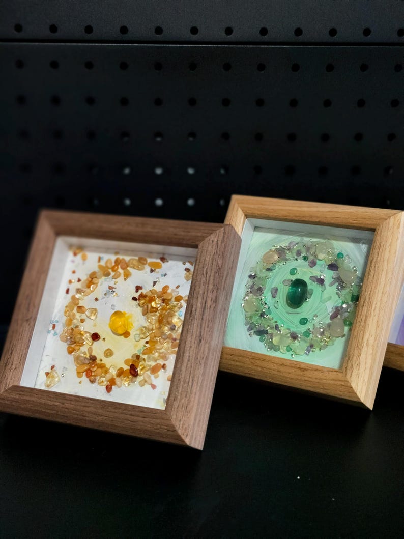 Pu&ograve; includere: Due opere d'arte quadrate incorniciate. Una presenta una pietra gialla centrale circondata da pietre arancioni e gialle su sfondo bianco. L'altra ha una pietra verde centrale con pietre verdi e viola su sfondo verde chiaro.