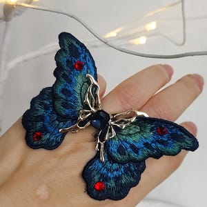 Geborduurde ring met vlindermotief, vleugels met een kleurverloop in turquoiseblauw, opvallende sieraden met strass-steentjes.