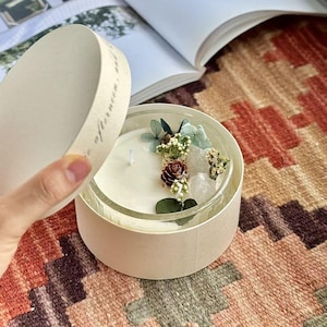 Peut inclure: Une bougie blanche dans un pot en verre transparent, placée dans une boîte ronde en carton beige avec un couvercle assorti. La surface de la bougie est décorée de feuilles vertes séchées, de petites fleurs blanches, d'une pomme de pin brune et de pierres transparentes.