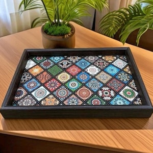 May include: A decorative wooden tray with a dark brown frame. The tray's interior features a colorful mosaic pattern of square tiles with intricate designs in various colors, including blue, orange, green, and red. The tray is placed on a wooden table.