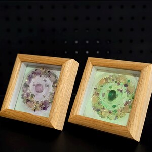 Pu&ograve; includere: Due opere d'arte quadrate incorniciate con cornici in legno chiaro. Una presenta una gemma viola al centro, circondata da pietre viola e trasparenti. L'altra ha una gemma verde al centro con pietre verdi e gialle.