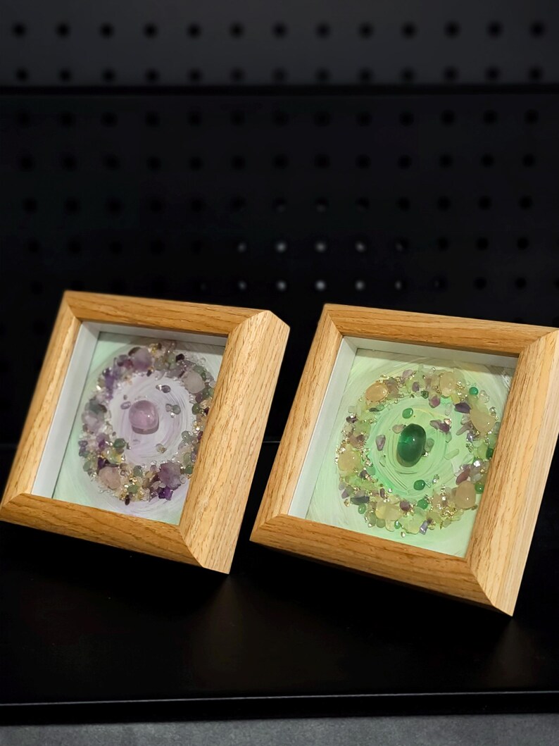 Pu&ograve; includere: Due opere d'arte quadrate incorniciate con cornici in legno chiaro. Una presenta una gemma viola al centro, circondata da pietre viola e trasparenti. L'altra ha una gemma verde al centro con pietre verdi e gialle.