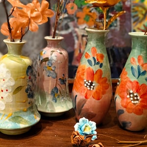 Puede incluir: Cuatro jarrones de cerámica pintados a mano con diseños florales. Los jarrones son en tonos verdes, rosas y naranjas, con detalles blancos y azules. Cada jarrón contiene flores secas. Los jarrones se exhiben sobre una superficie de madera.