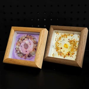 Pu&ograve; includere: Due opere d'arte incorniciate con cornici in legno. Una presenta un design viola e rosa con cristalli, l'altra ha una disposizione di cristalli arancioni e gialli. Entrambe sono quadrate e poste su uno sfondo scuro.